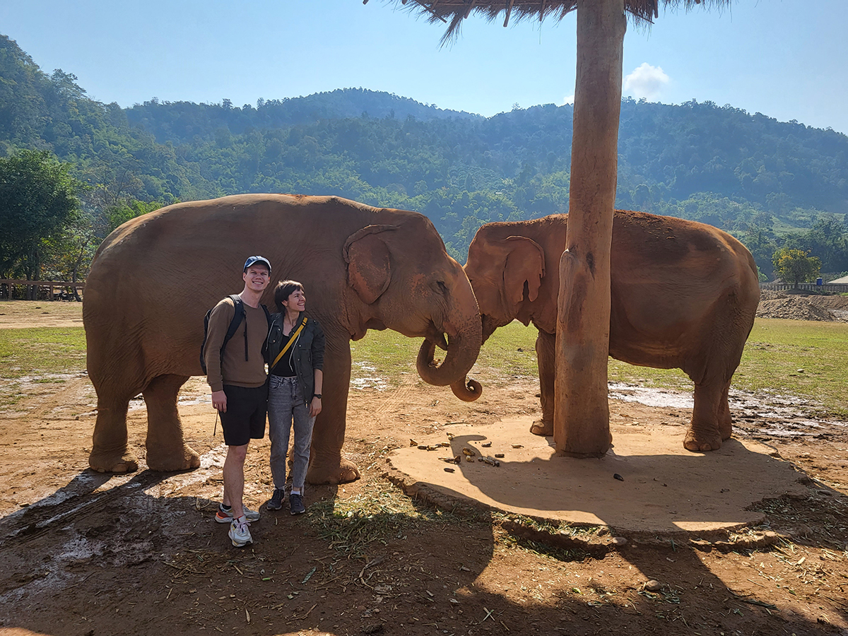 Thailand's Elephant Nature Park A place full of love, respect and bananas Ein Paar steht neben zwei Elefanten auf einer Safari mit Berge im Hintergrund.