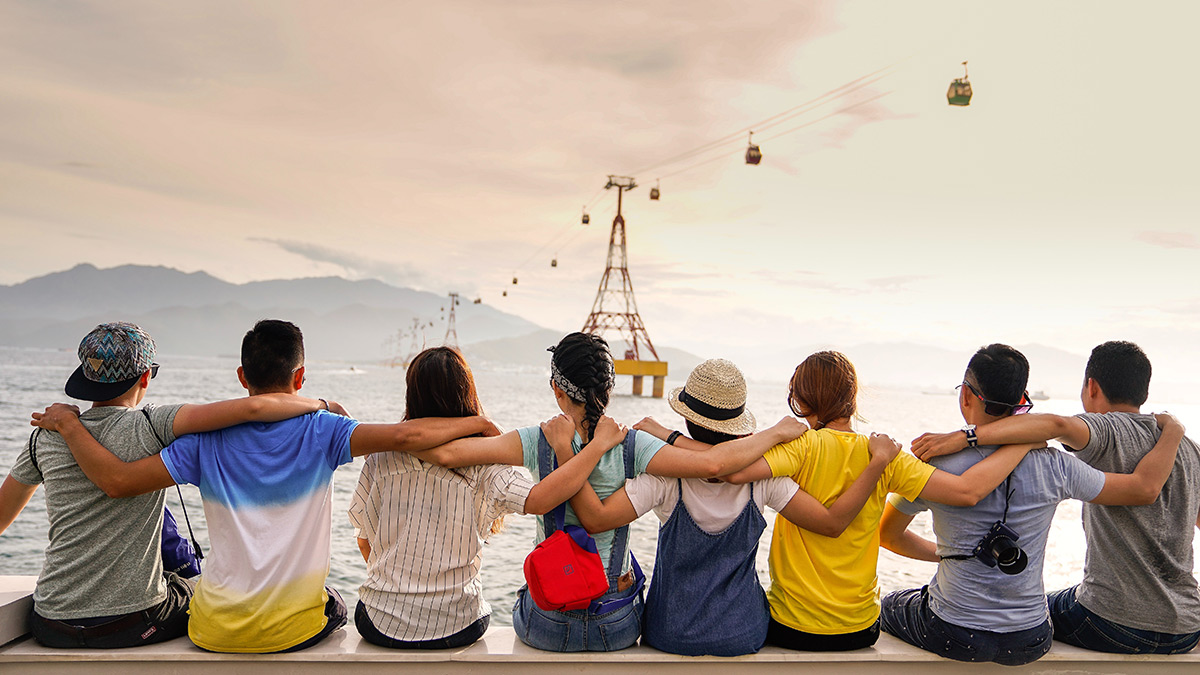 Group travel for solo travelers: Tips, destinations & benefits Eine Gruppe von sieben Freunden sitzt zusammen am Wasser und blickt auf eine Seilbahn über dem Meer.