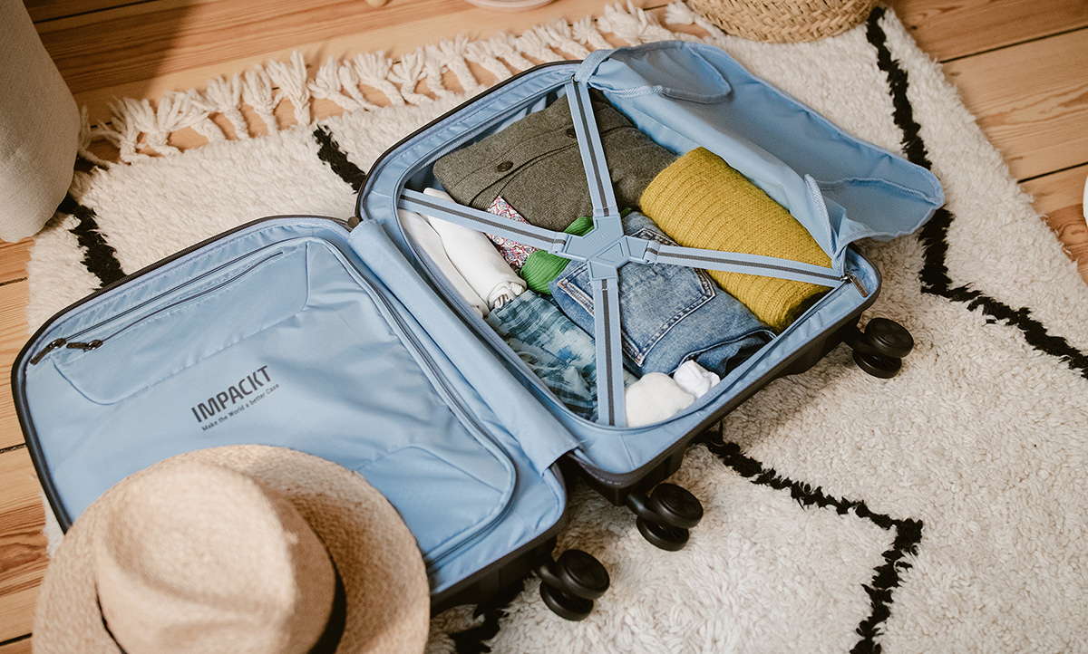 Packing your suitcase - but smart and sustainable please Ein geöffneter blauer Koffer, gepackt mit Kleidung und Reisverschlüssen, liegt auf einem hellen Teppich in einem wohnlichen Raum. Neben dem Koffer liegt ein Strohhut.