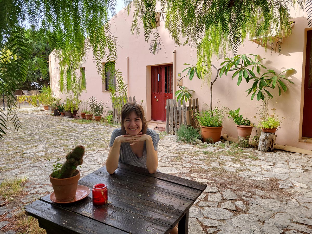Eco-friendly road trip in Sicily: Franzi's travel adventure with the hybrid car Lächelnde Frau an Tisch mit Kaktus und rotem Windlicht, umgeben von Pflanzen und rosafarbenem Gebäude im Hintergrund
