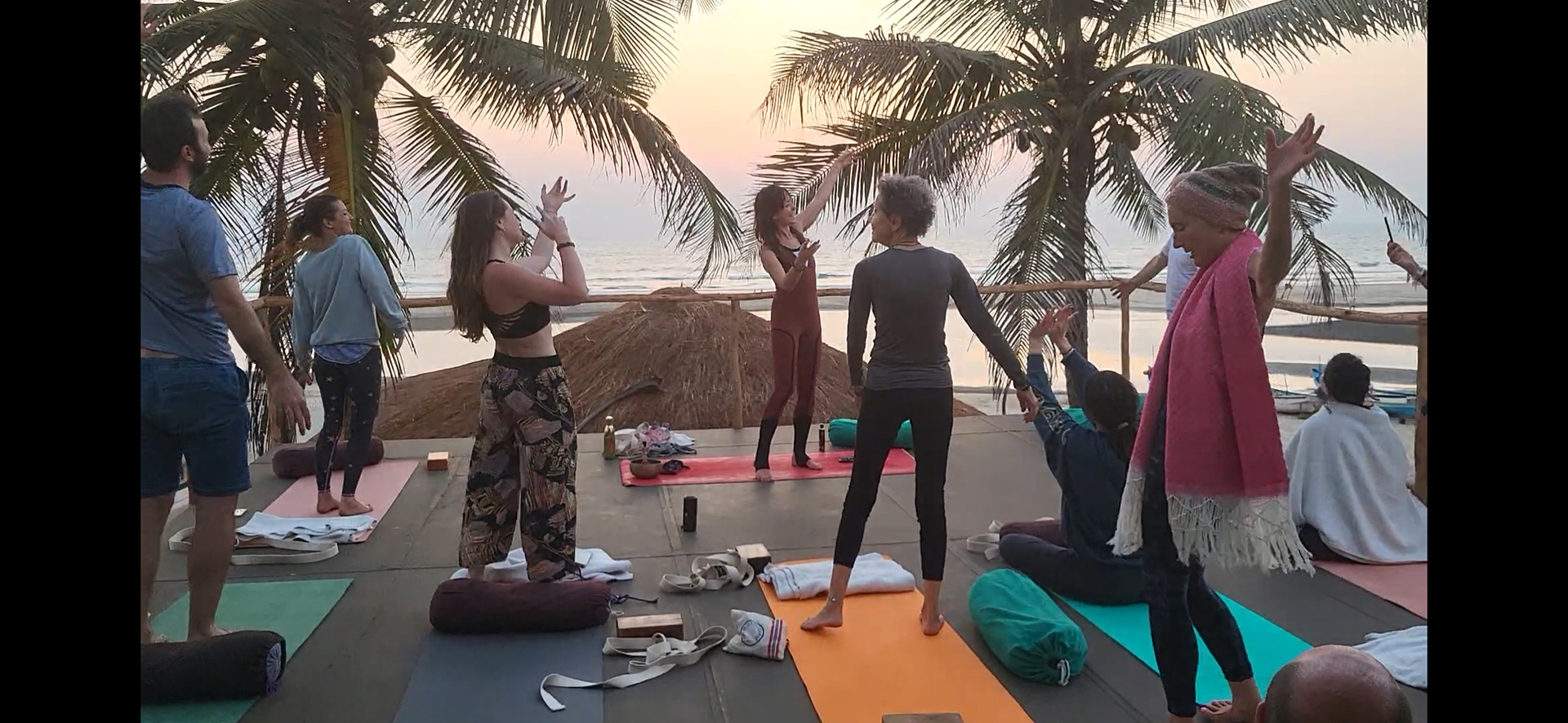Yoga, beach and sustainability: my perfect detox holiday in India Menschen praktizieren Yoga in einer Gruppe auf einer Terrasse mit Meerblick und Palmen im Hintergrund bei Sonnenuntergang.