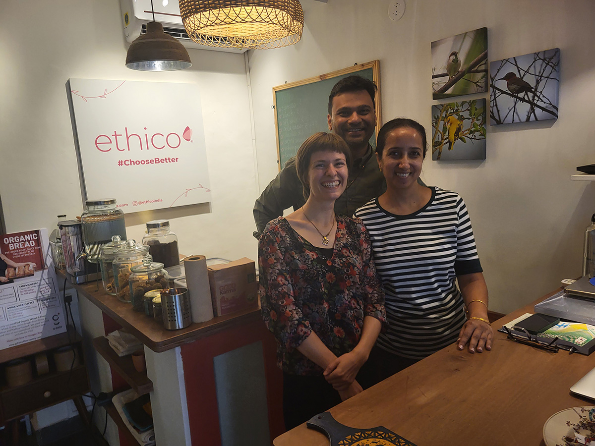 Sustainable travel in Goa: A visit to the zero-waste store Ethico and the initiative "Offbeat in Goa" Drei Personen stehen hinter der Theke in einem Café mit ethischem Schwerpunkt, lächeln in die Kamera. Im Hintergrund sind Schilder und Getränke zu sehen.