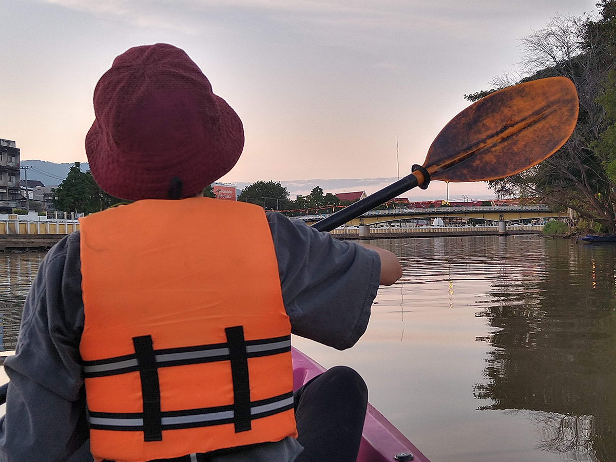 Müll sammeln bei romantischer Kajak-Tour: eine Aktion der besonderen Art Rückansicht einer Person mit orangefarbener Schwimmweste und rotbraunem Hut, beim Paddeln in einem Kajak auf einem Fluss bei Sonnenuntergang