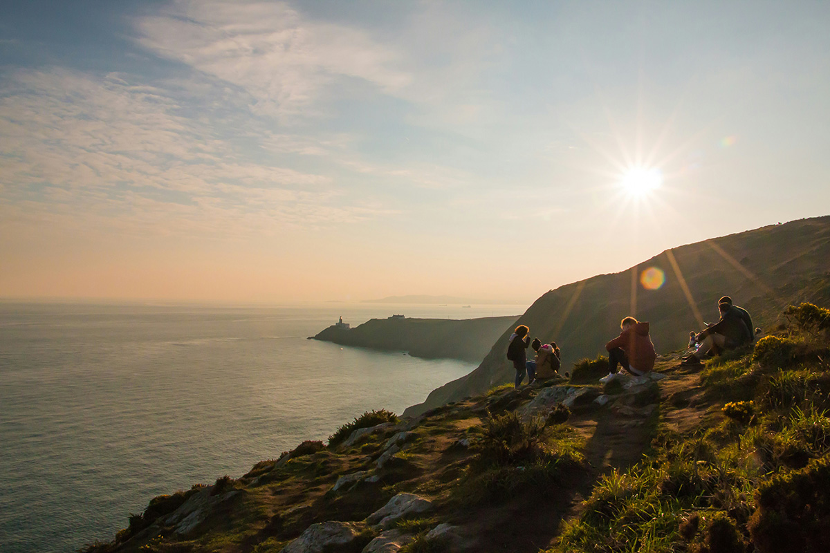 Die 4 schönsten Glücksorte für Mensch und Umwelt Menschen sitzen auf einem Hügel mit Blick auf das Meer und einen Leuchtturm im Sonnenuntergang