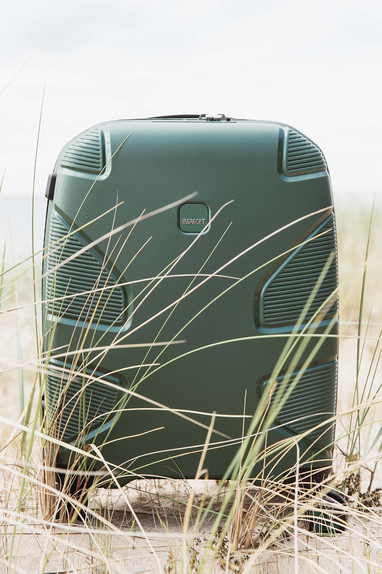 Robuster grüner Hartschalenkoffer am Strand Grüner Hartschalenkoffer steht auf sandigem Boden zwischen Dünengras am Strand.