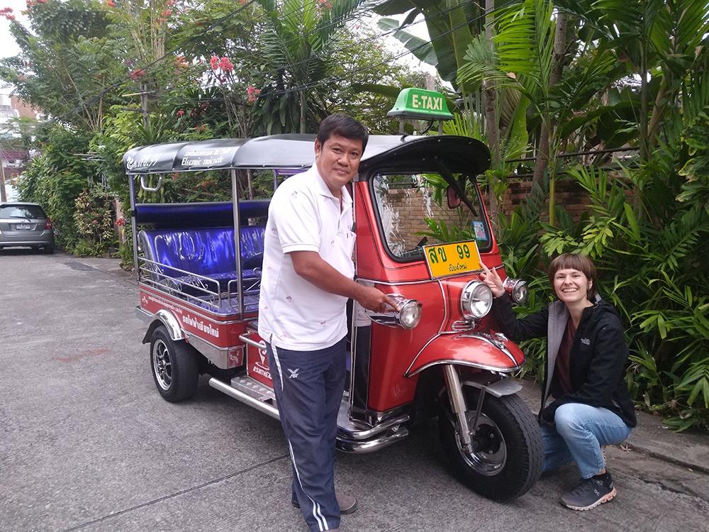 Frischluft-Abenteuer in Thailand: Entdecke Chiang Mai im grünen E-Tuk-Tuk Ein Mann und eine Frau lächeln neben einem roten Elektro-Tuk-Tuk mit grünem E-Taxi-Schild, umgeben von üppigen Pflanzen