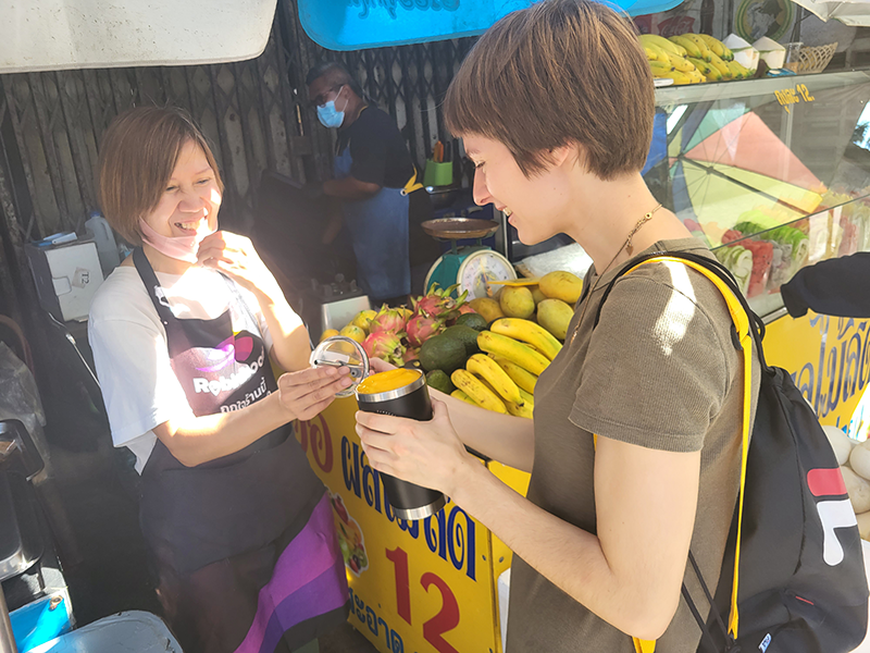 Thailand, the vegan paradise? - Local restaurants and specialities you should try Zwei Frauen lächeln an einem Obststand, während eine von ihnen einen Smoothie hält.