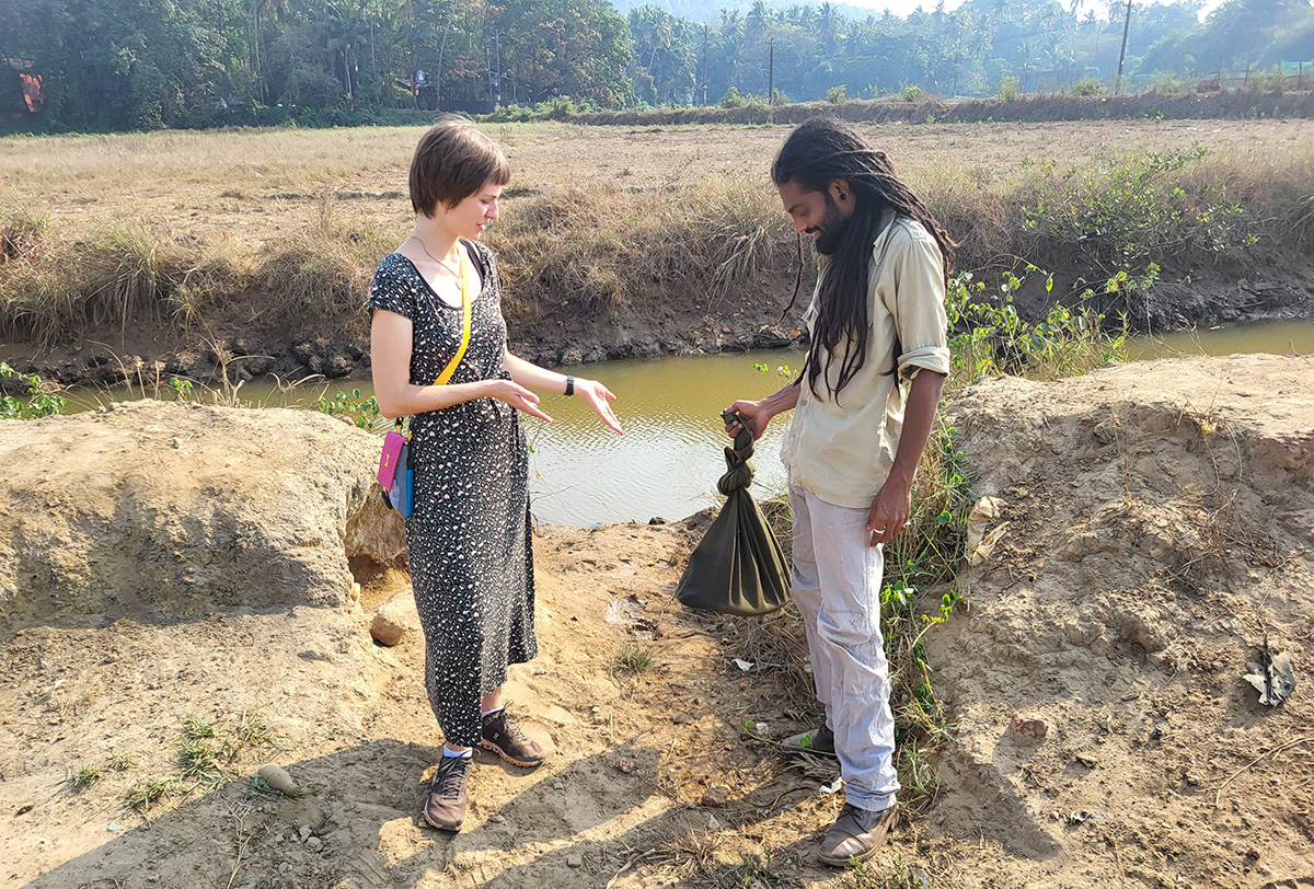 Snake whisperer in India: Between poison fangs and climbing monkeys Eine Frau und ein Mann stehen an einem Flussufer und unterhalten sich. Der Mann hält einen gefüllten Stoffbeutel in der Hand.