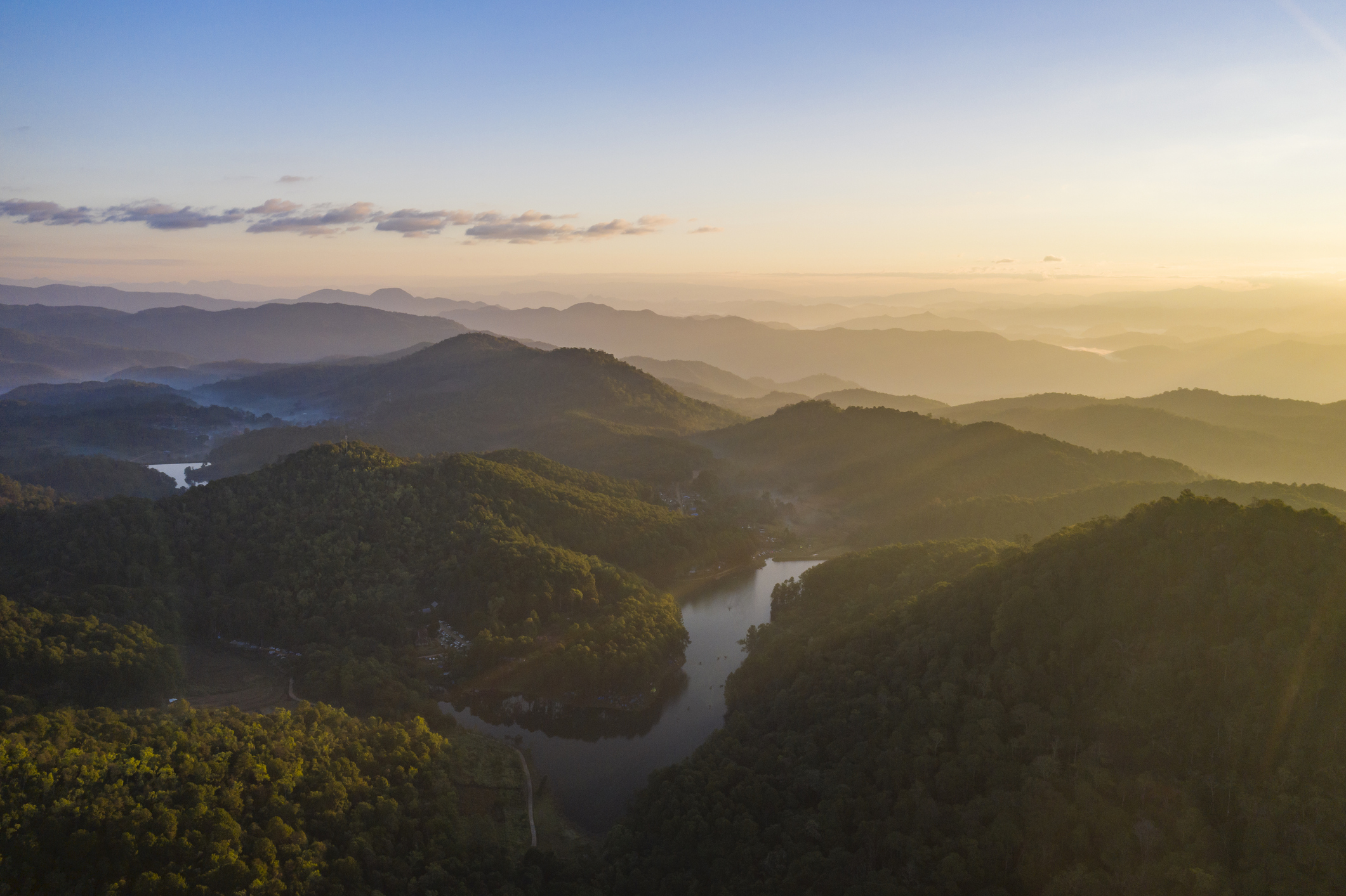 Luftaufnahme einer malerischen Hügellandschaft bei Sonnenaufgang Malerische Hügellandschaft bei Sonnenaufgang mit sanften, bewaldeten Hügeln und einem ruhigen Fluss, aufgenommen aus der Luft.