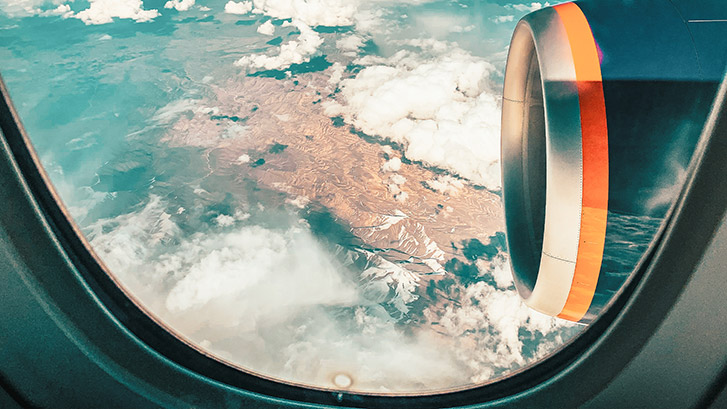 Blick aus dem Flugzeugfenster auf Landschaft und Wolken Fotografie durch ein Flugzeugfenster mit Sicht auf eine Landschaft mit Bergen und Wolken sowie einem Triebwerk im Vordergrund