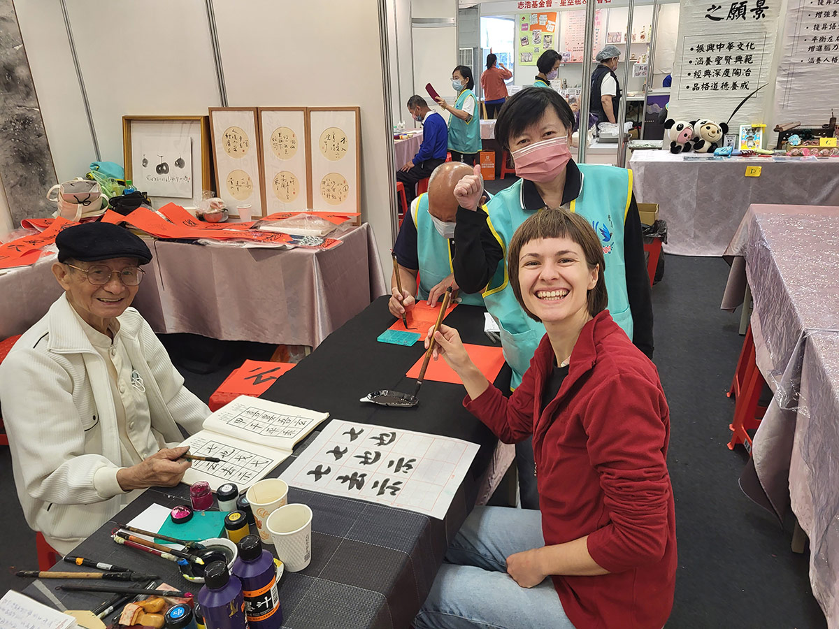 Enchanting calligraphy art in Taiwan Lächelnde Teilnehmer bei einem Kalligraphie-Workshop auf einer Messe, umgeben von Materialien und Kunstwerken.
