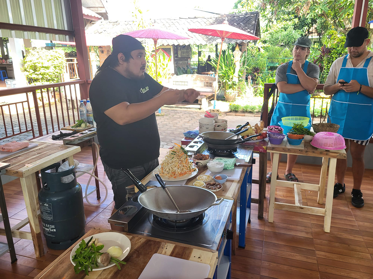 Traditional Thai cooking course on an organic farm? - Pretty spicy. Ein Koch gibt einen Kochkurs im Freien, umgeben von Zutaten wie Gemüse und Gewürzen, während zwei Teilnehmer zuschauen.