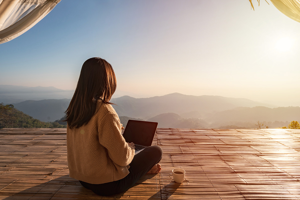 Workation: Alles übers Arbeiten mit Urlaubsfeeling Eine Frau sitzt auf einer Terrasse mit Laptop und Kaffee, Blick auf die Berge und Sonnenaufgang