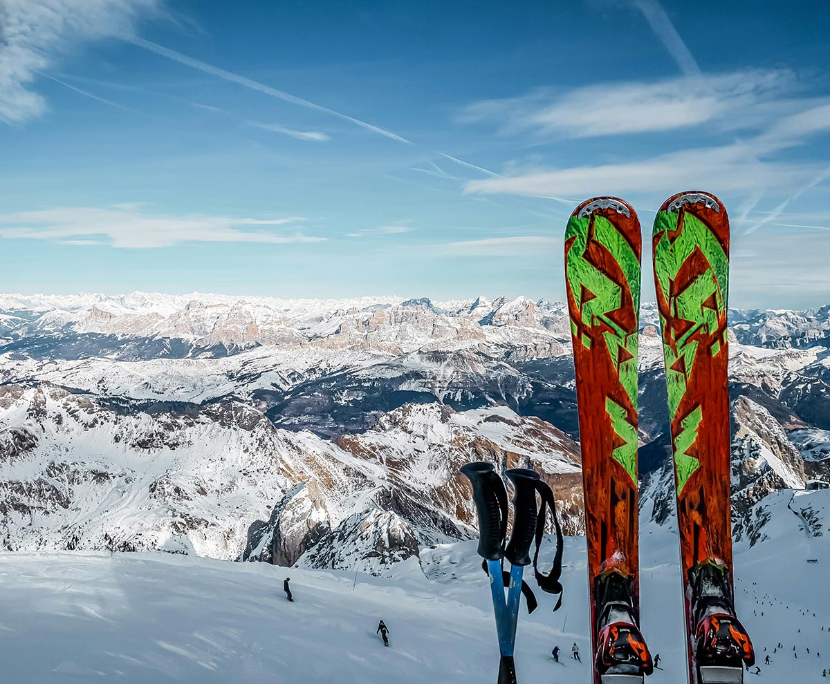 Ski vacation packing list: what not to miss Ein Paar bunte Skier und Skistöcke auf einem schneebedeckten Berg mit einer atemberaubenden Aussicht auf die Alpen im Hintergrund.