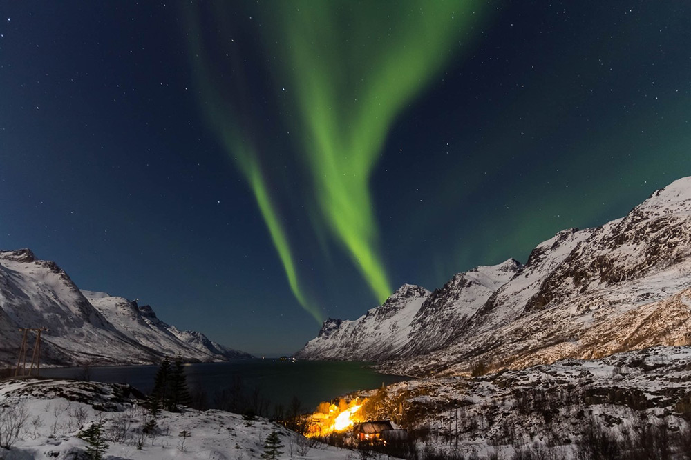 Nordlichter über verschneiten Bergen und beleuchtetem Tal Grünes Nordlicht erhellt den Nachthimmel über einer verschneiten Gebirgslandschaft mit einem hell erleuchteten Tal.