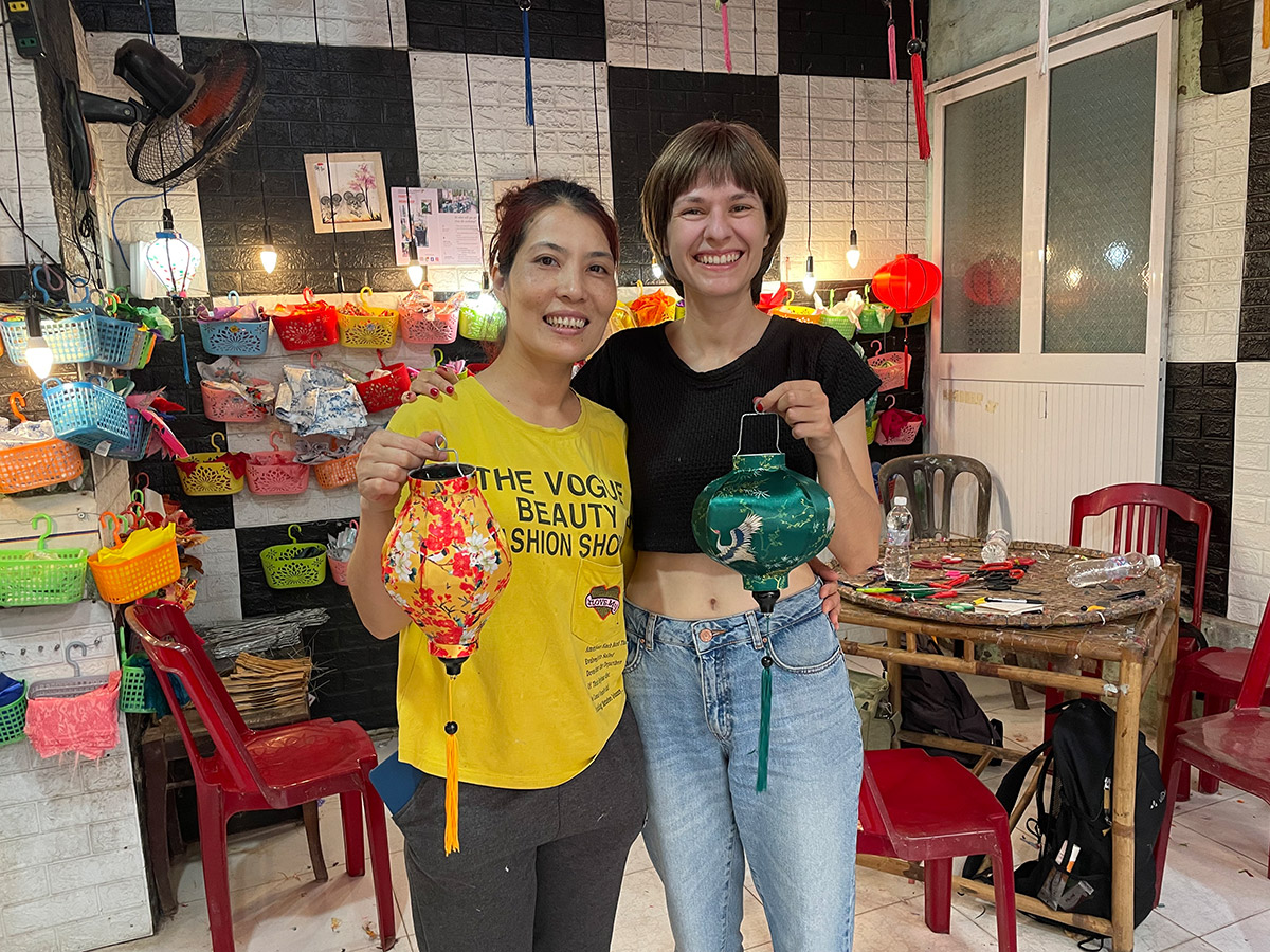 Unique craft workshop in Hoi An: Create your own lantern with the Lantern Lady  Zwei Frauen lächeln und halten bunte Laternen in einem Handwerksladen, umgeben von farbenfrohen Körben und Bastelmaterialien.