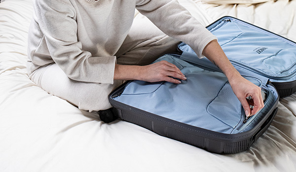 Person packt einen Koffer auf einem Bett Eine Person in beiger Kleidung sitzt auf einem Bett und packt einen blauen Koffer. Der Koffer ist offen und die Person schließt den Reißverschluss.
