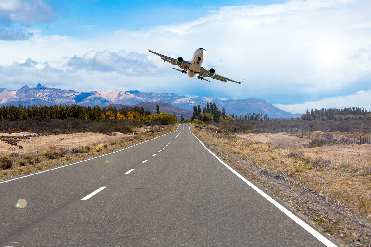 Offset your flight: Your steps to CO2 offsetting Ein Flugzeug hebt über einer langen, geraden Landstraße ab. Im Hintergrund sind Berge und bewaldete Flächen zu sehen.