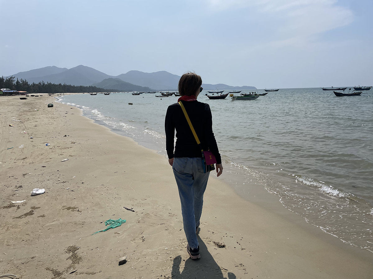 Garbage on the horizon: How plastic mountains changed my personal journey Eine Person geht am Strand entlang, während Boote im Meer treiben und Berge im Hintergrund zu sehen sind.
