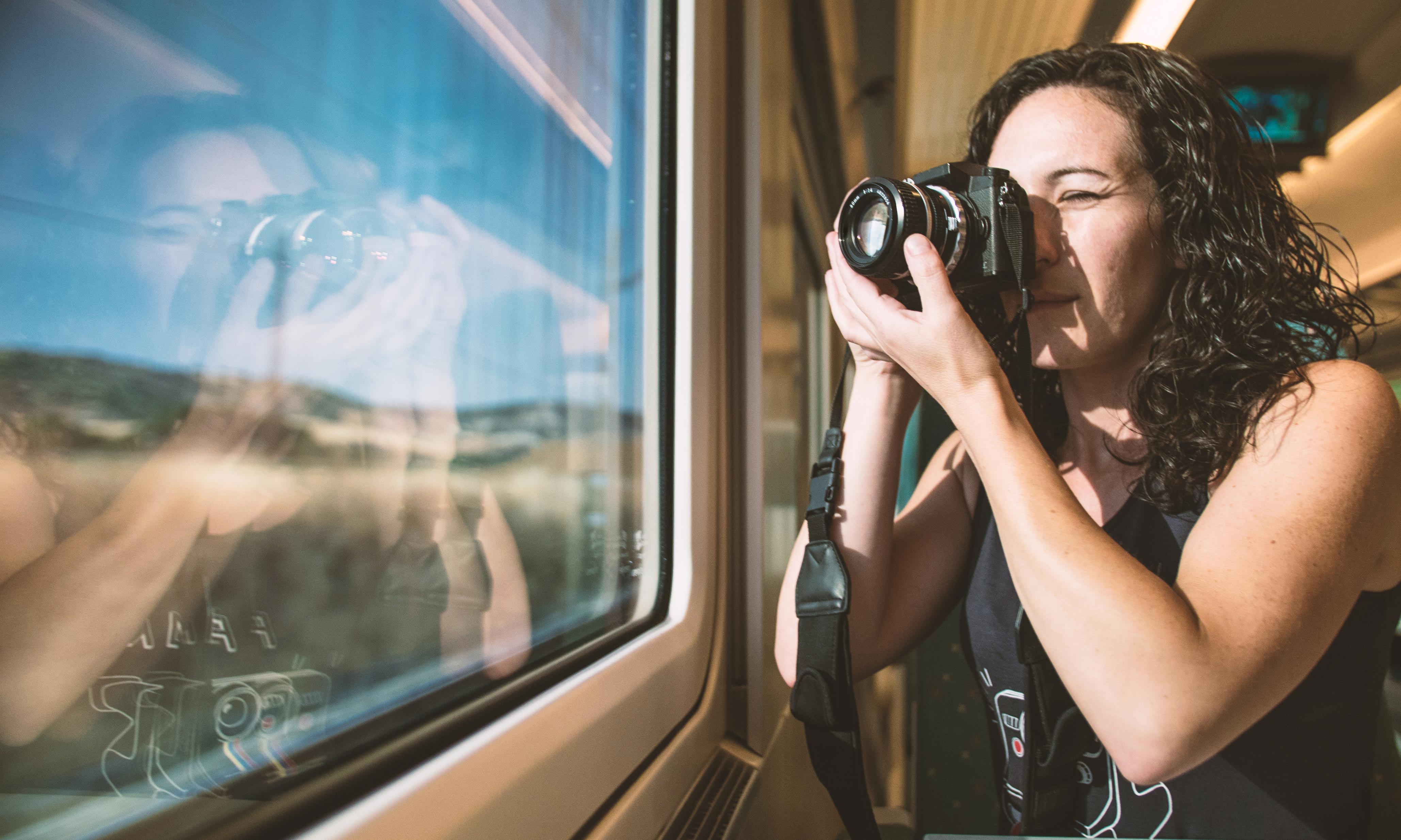 Sustainable travel - be smart and start your vacation more responsibly with IMPACKT Eine Frau mit lockigen Haaren macht mit einer Kamera ein Foto aus dem Fenster eines fahrenden Zuges. Ihre Reflexion ist im Glas sichtbar, während die Landschaft vorbeizieht.