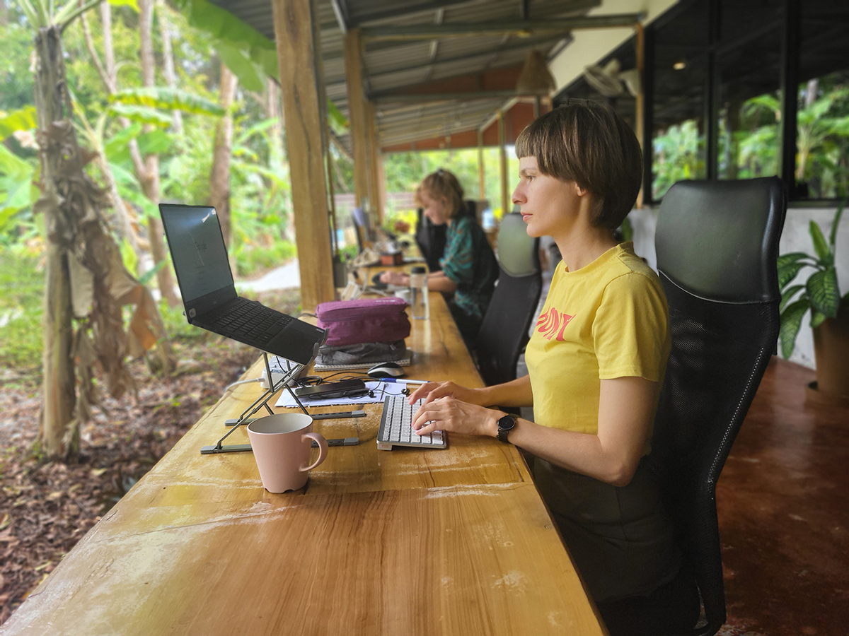 Workation as a paradisiacal working model: Home office under palm trees, end of work on the beach Zwei Personen arbeiten an Laptops in einem offenen Büro im Grünen, umgeben von Pflanzen, mit Kaffeetassen auf dem Tisch.