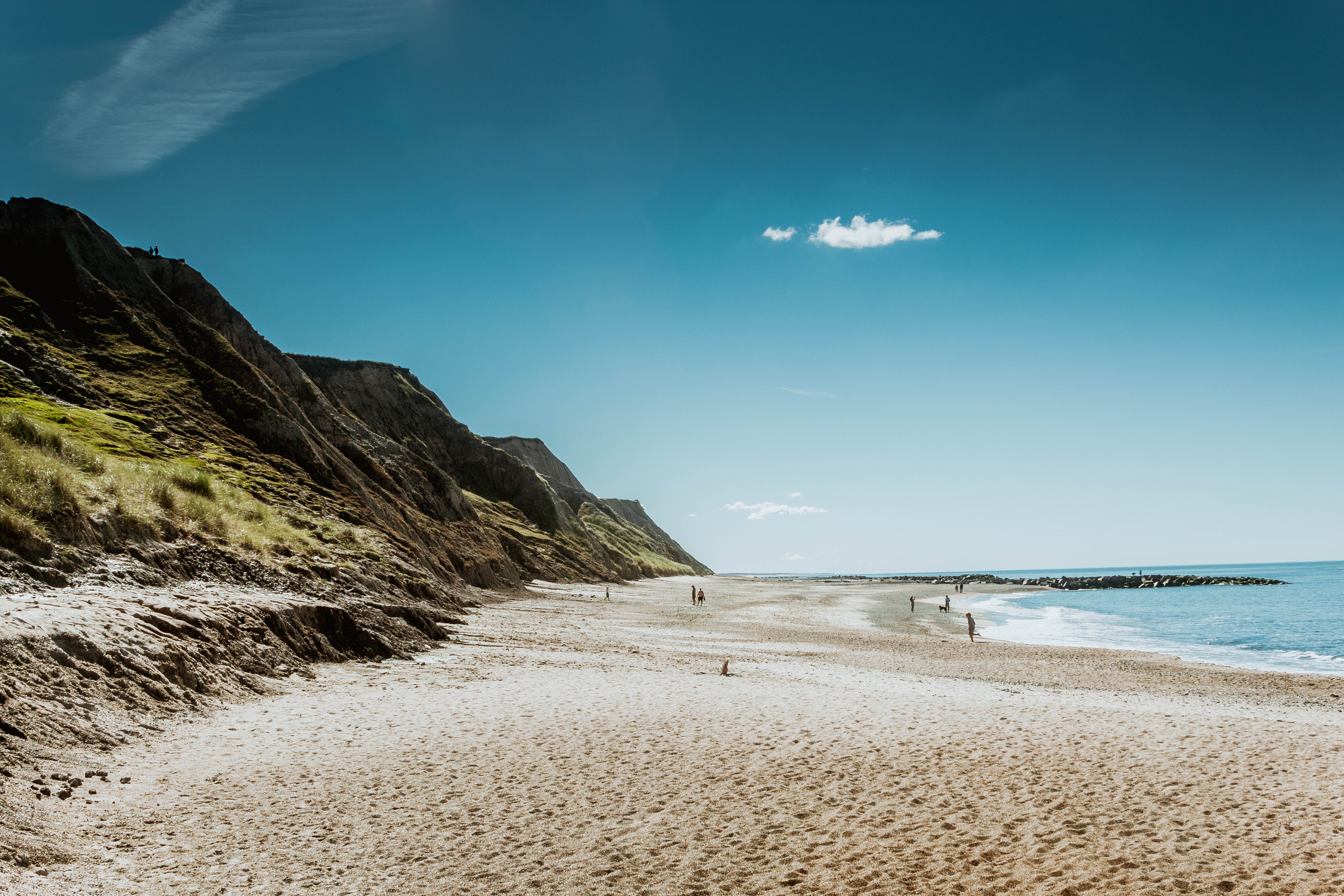 Idyllischer Strand mit Klippen und blauem Himmel Ein ruhiger Strand mit hohen Klippen, goldenem Sand und blauem Himmel, im Hintergrund einige Menschen im Wasser und am Ufer